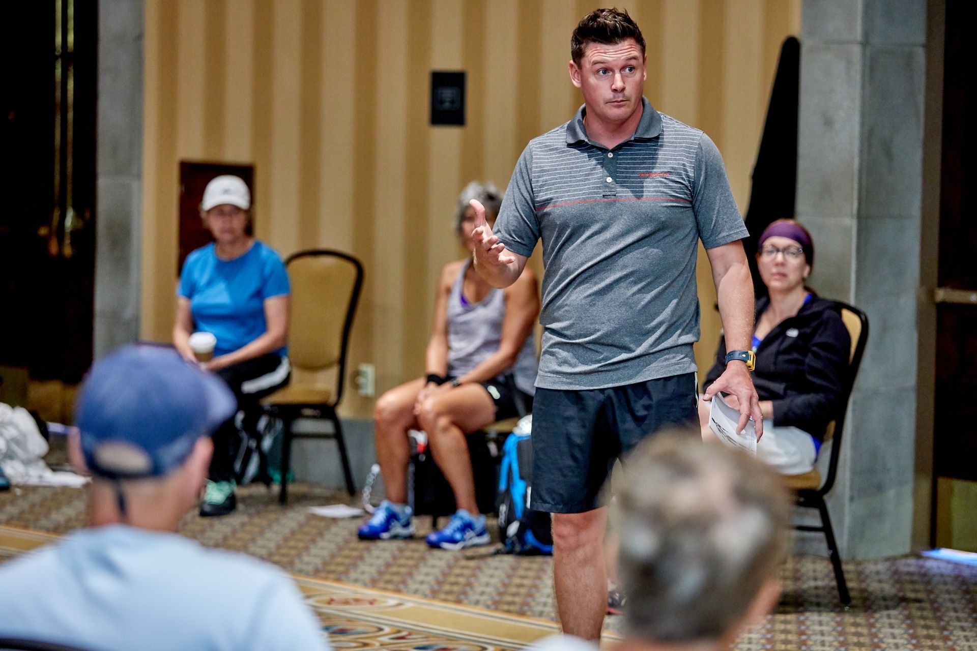 Man in gray shirt addresses a seated group; hotel conference room.