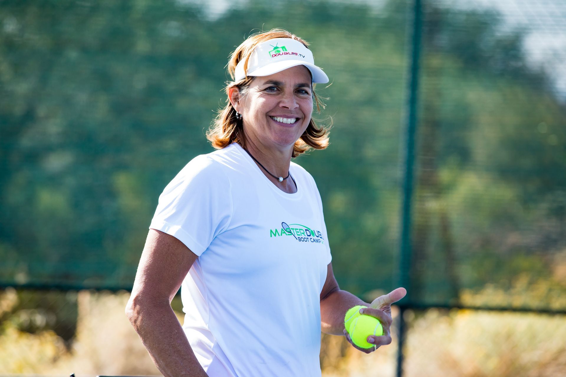 Woman in white shirt and visor holding a tennis ball, smiling outdoors on a tennis court.