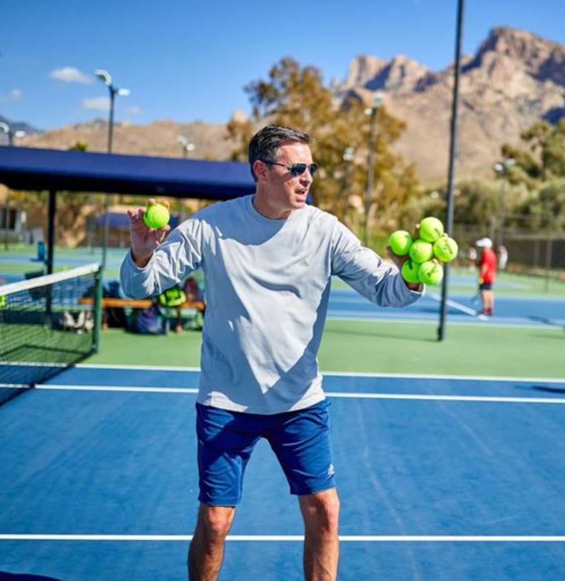 Man on a tennis court holding tennis balls. Blue court, mountain backdrop, sunny day.