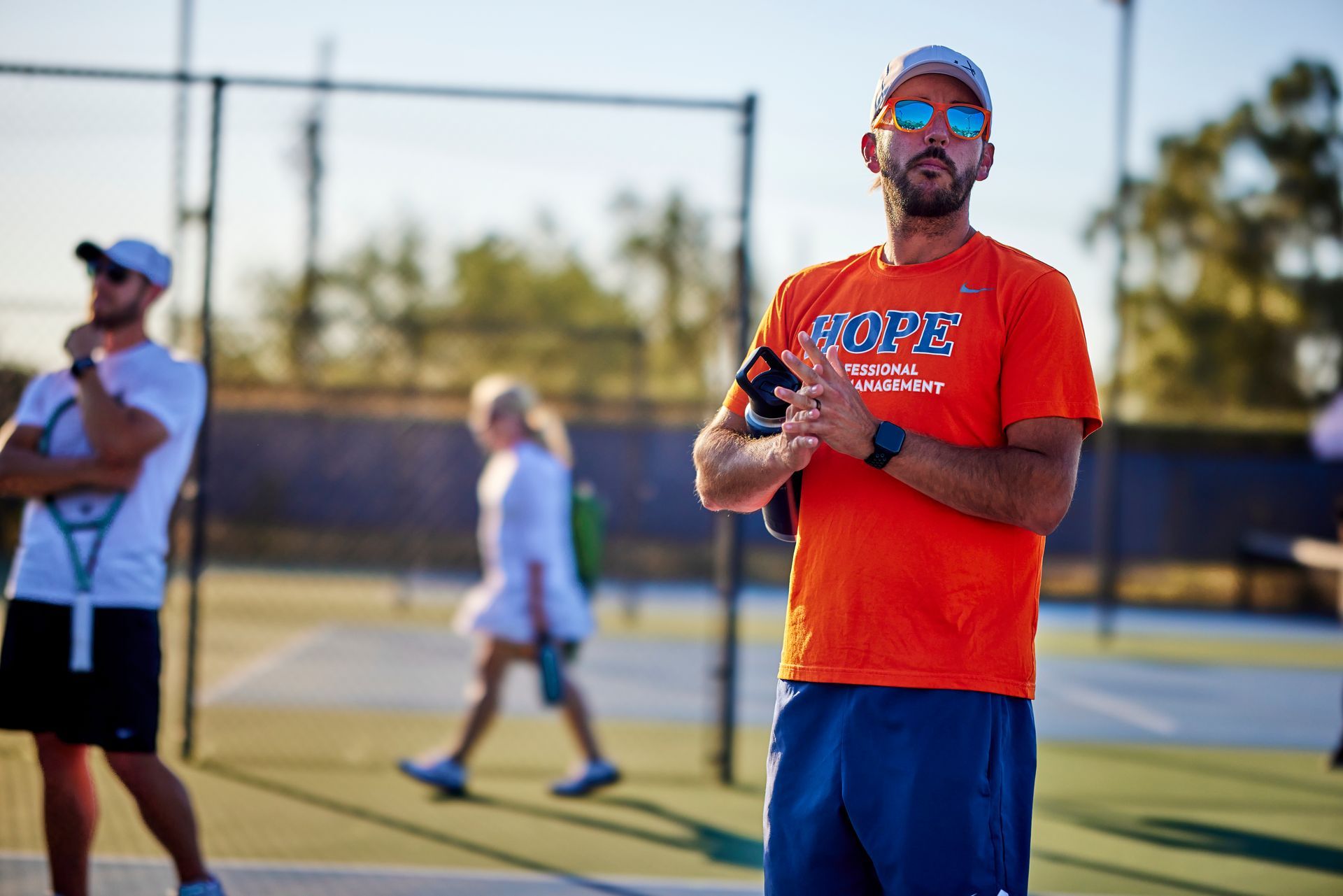 Man in orange shirt and blue shorts holding a drink on a tennis court, with players in the background