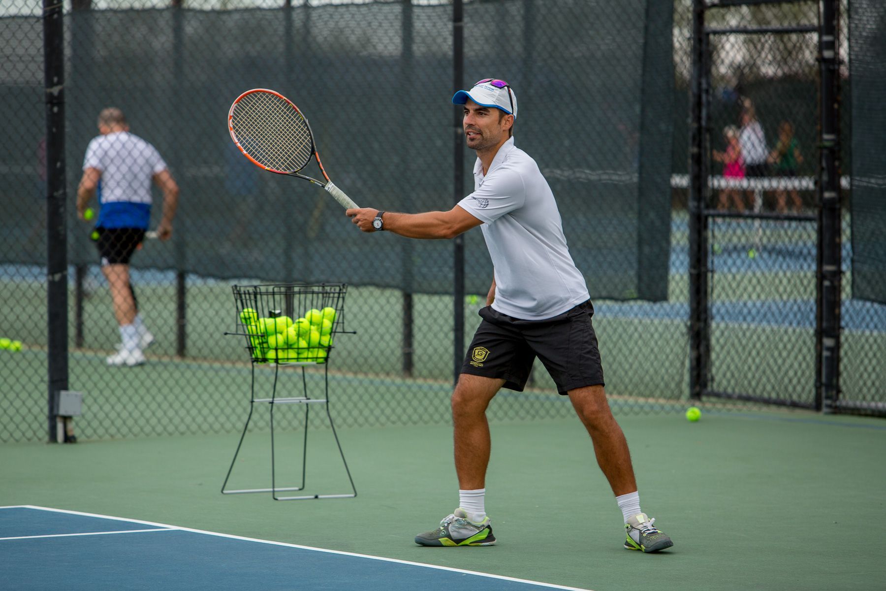 Tennis player on court, swinging racket; basket of balls nearby.