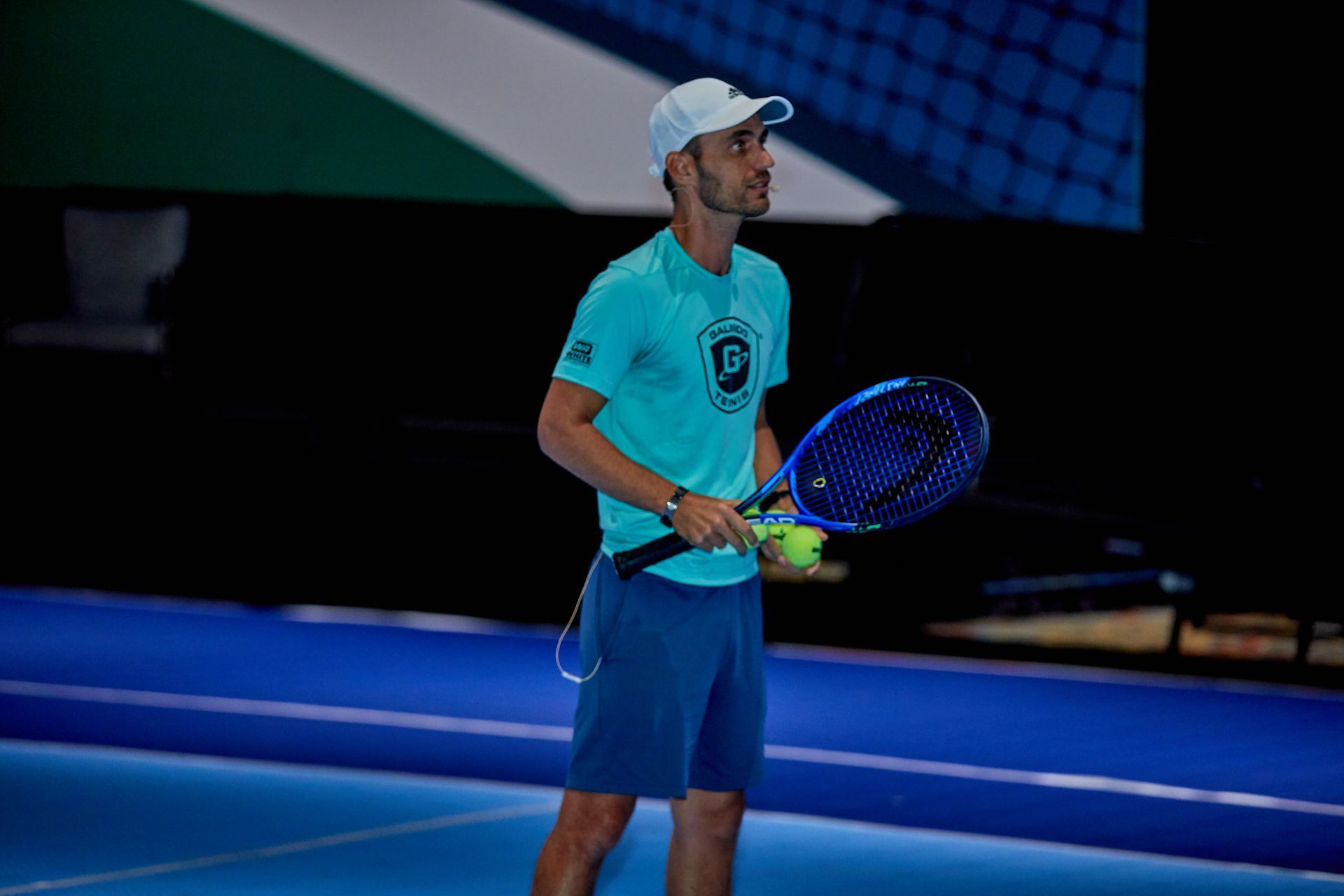 Tennis player holding racket and balls on a blue court. Wearing teal shirt, cap, and blue shorts.