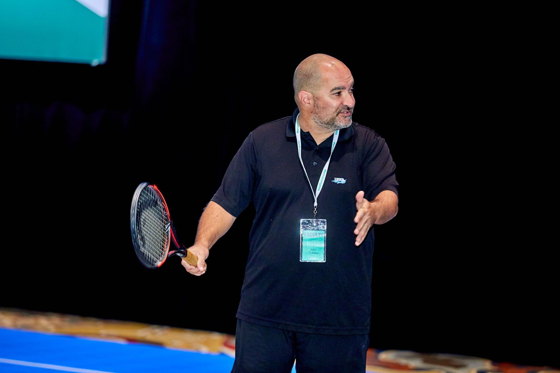 Man holding a tennis racket, gesturing with hands. Dark blue shirt, lanyard. Black background.