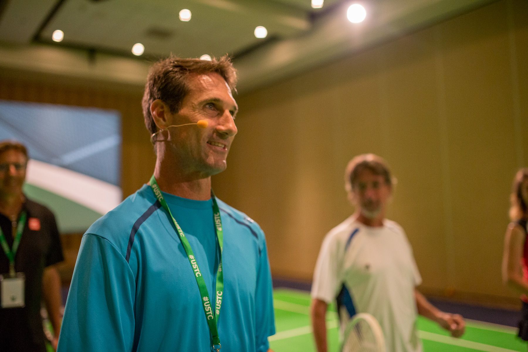 Man in blue shirt smiles, indoors. Others behind him; green lanyard, green floor.