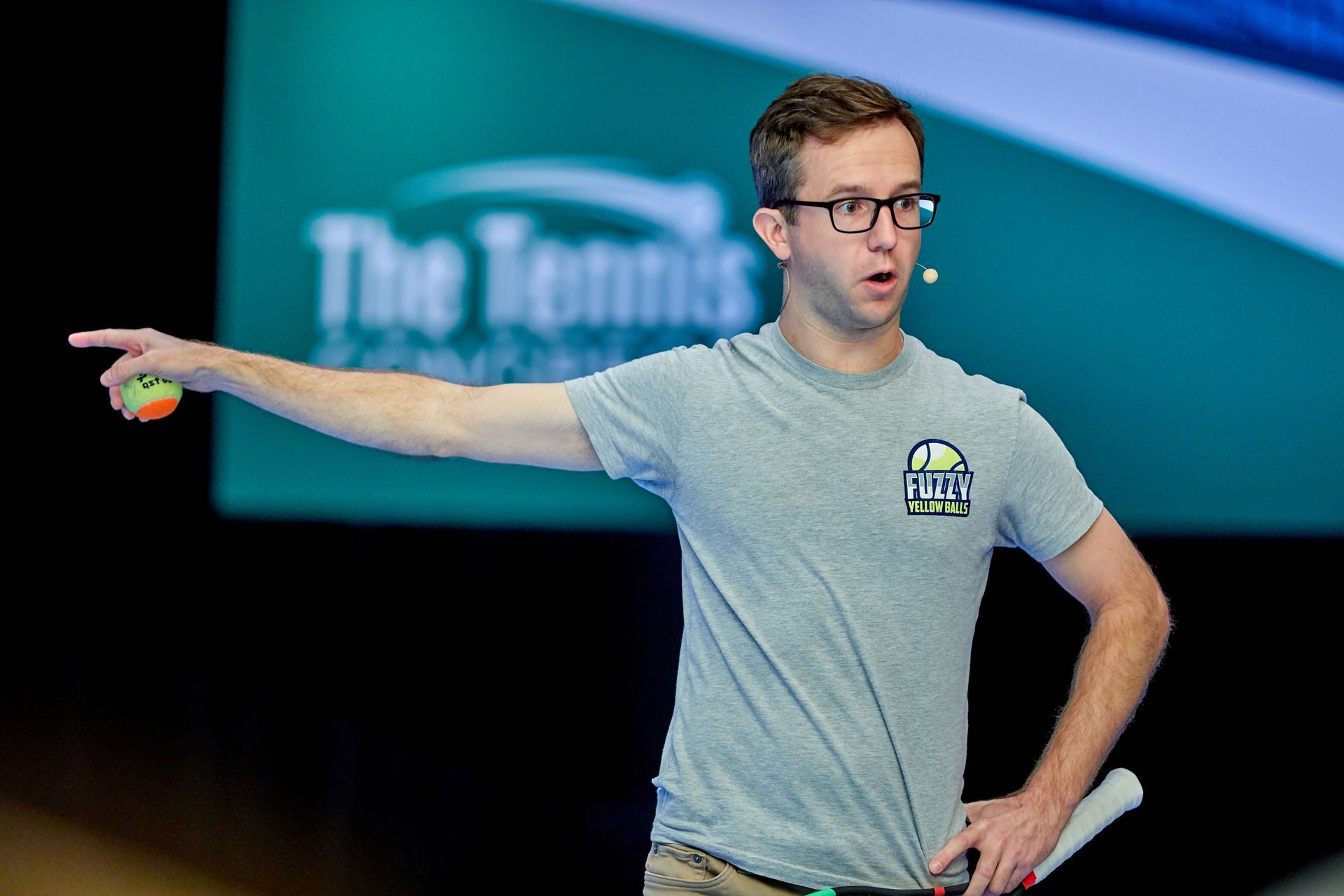 Man holding a tennis ball and racket, pointing left while speaking at a 