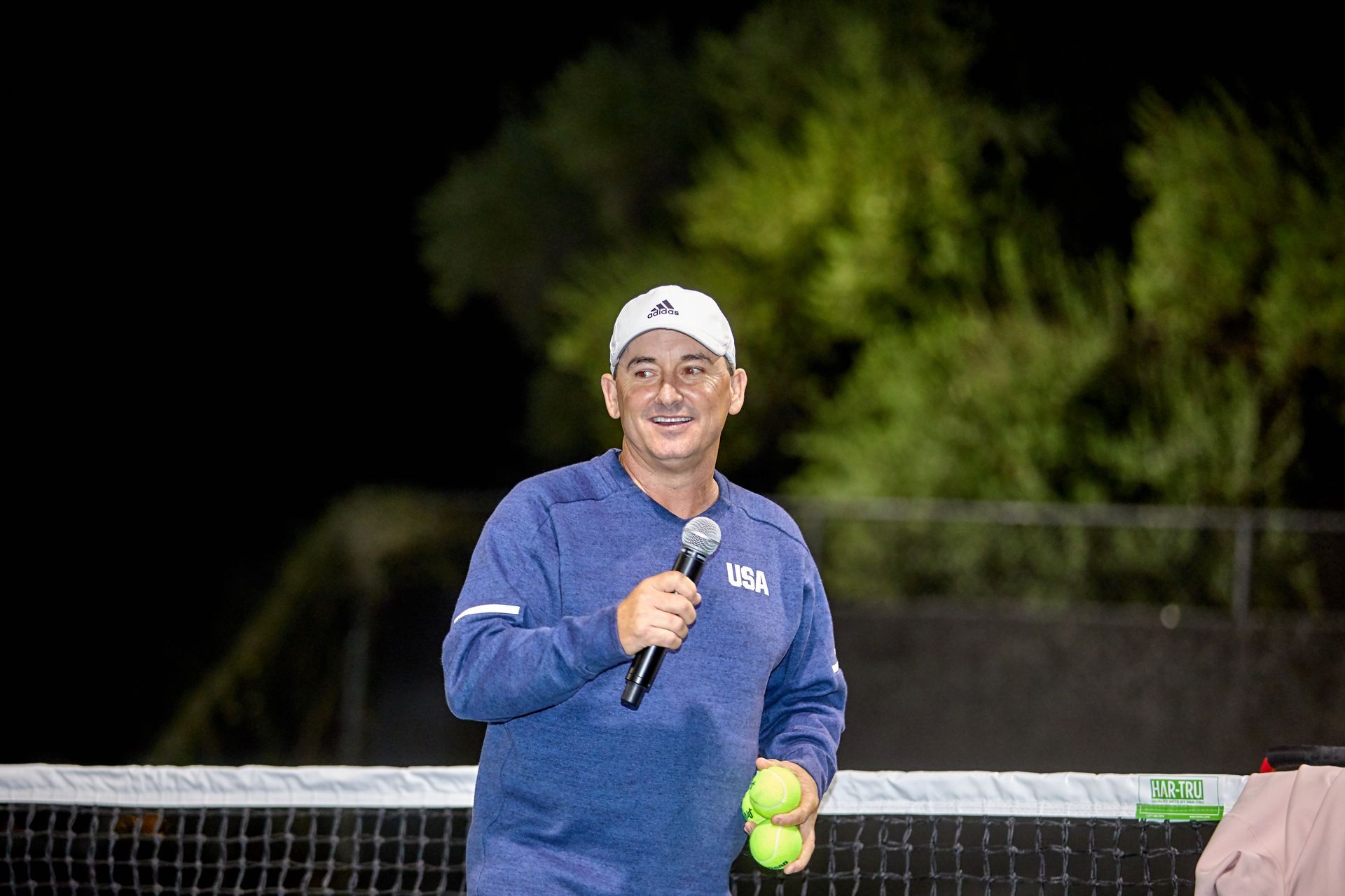 Man in blue sweater and white hat holding a microphone and tennis balls on a tennis court at night.
