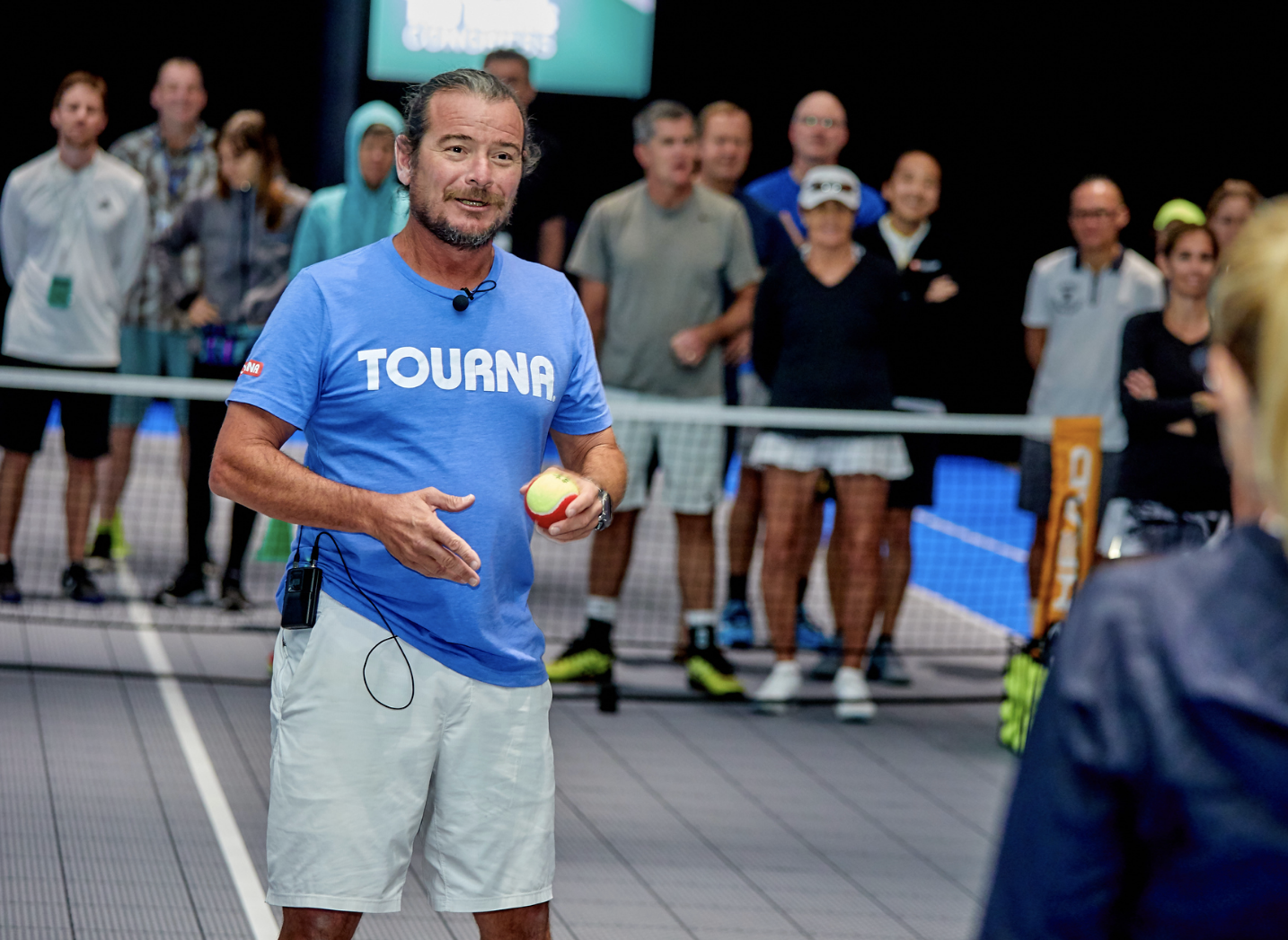 Man in blue shirt, holding a tennis ball, speaks to group near a tennis court.