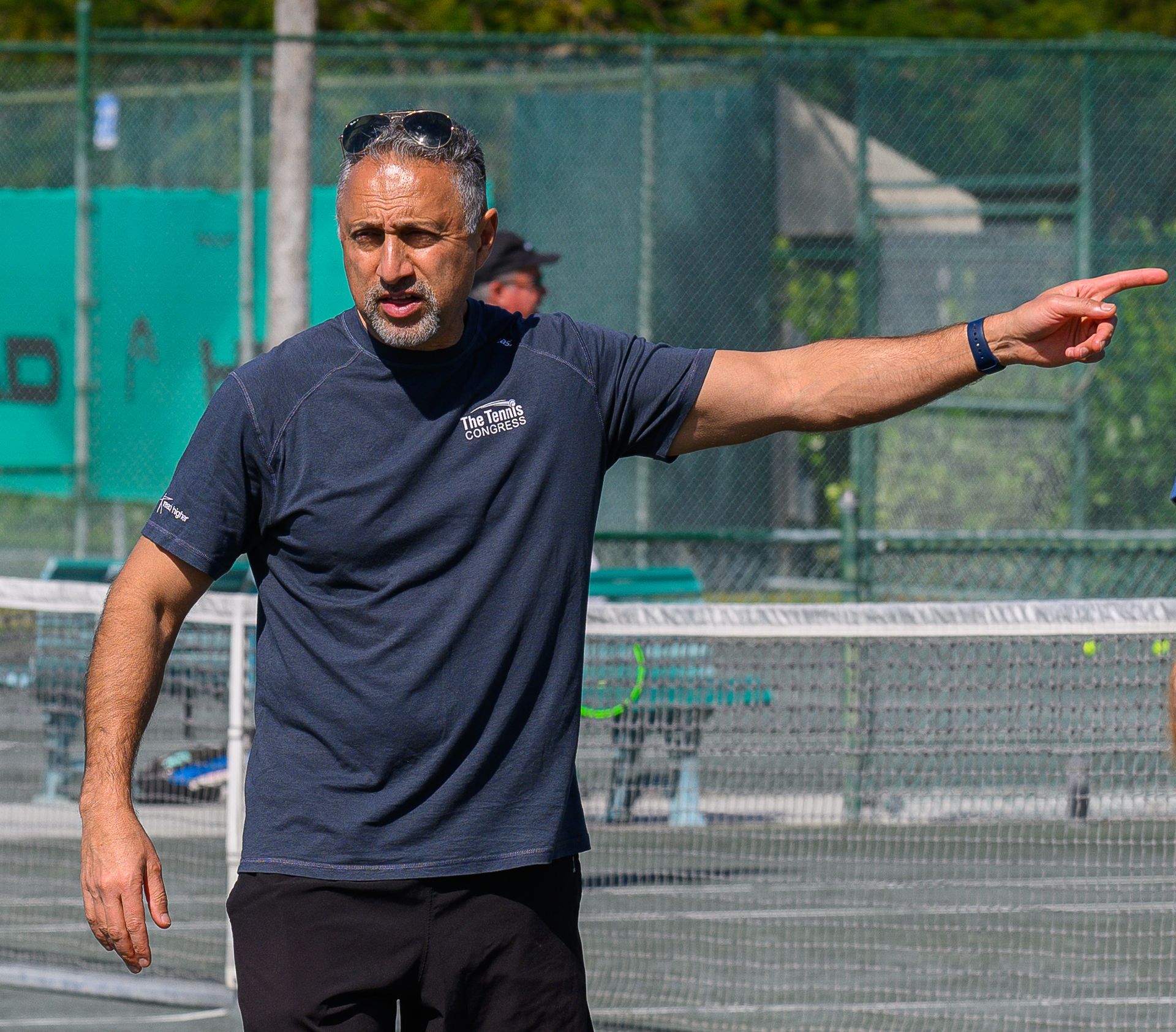 A coach in a dark blue shirt and sunglasses stands on a tennis court, gesturing to the side.
