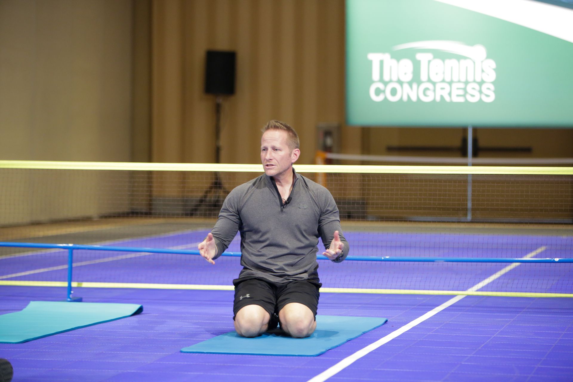 Man on a blue mat, gesturing with hands; tennis court setting with a 