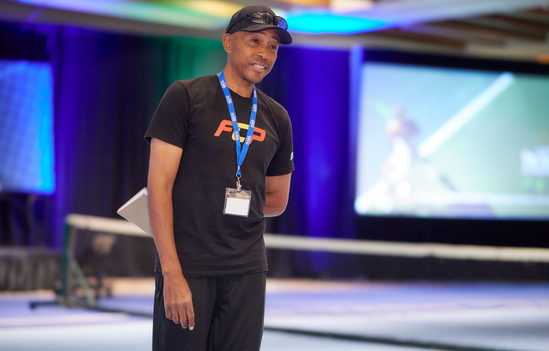 Man in black t-shirt and cap stands on stage, holding a clipboard, speaking during a presentation.