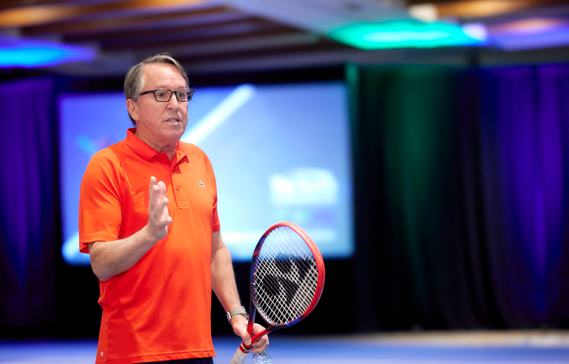 Man in orange shirt holds a paddle, gesturing while standing in a room with a screen.