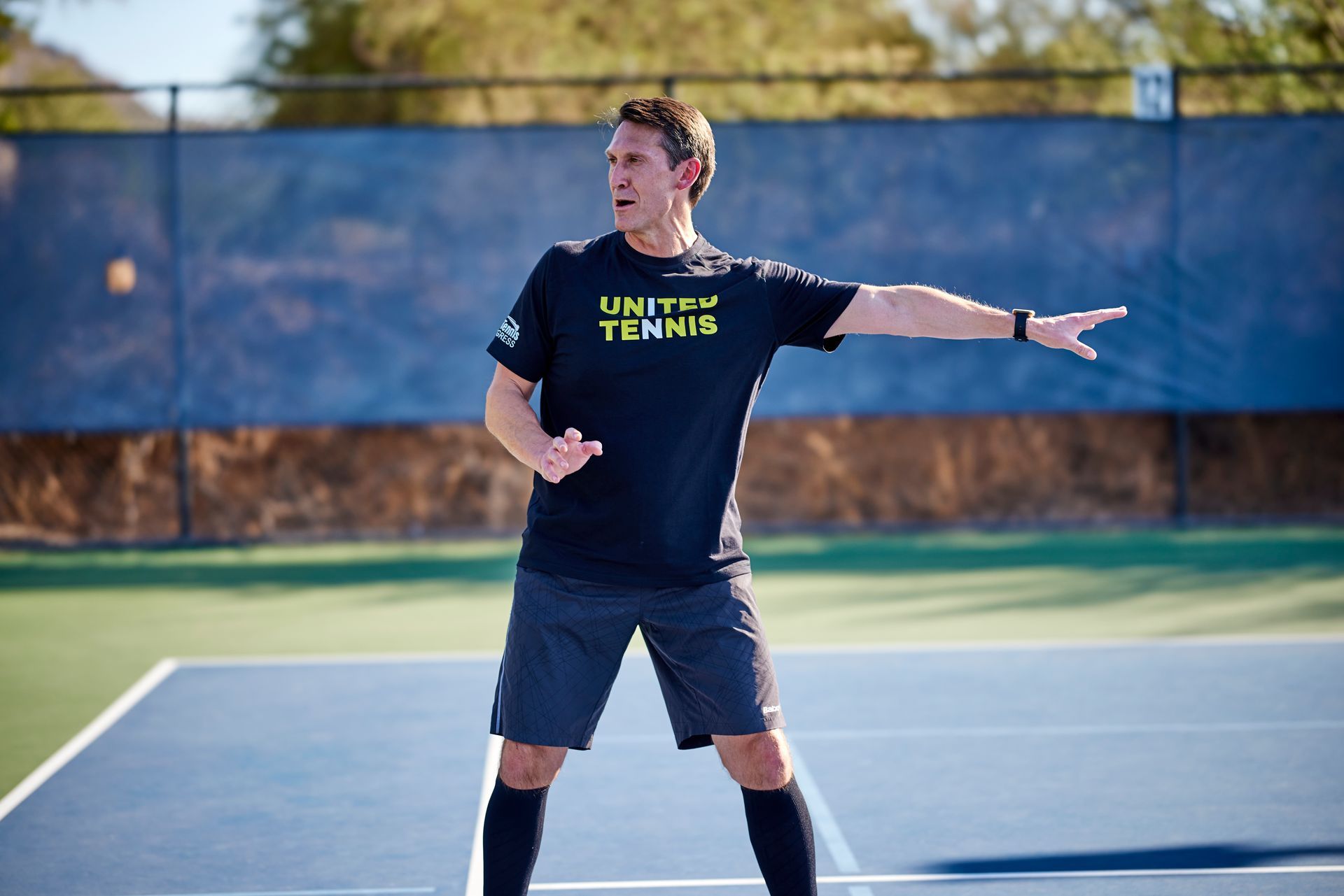 Man on tennis court in black shirt, shorts, and socks, gesturing with hands.
