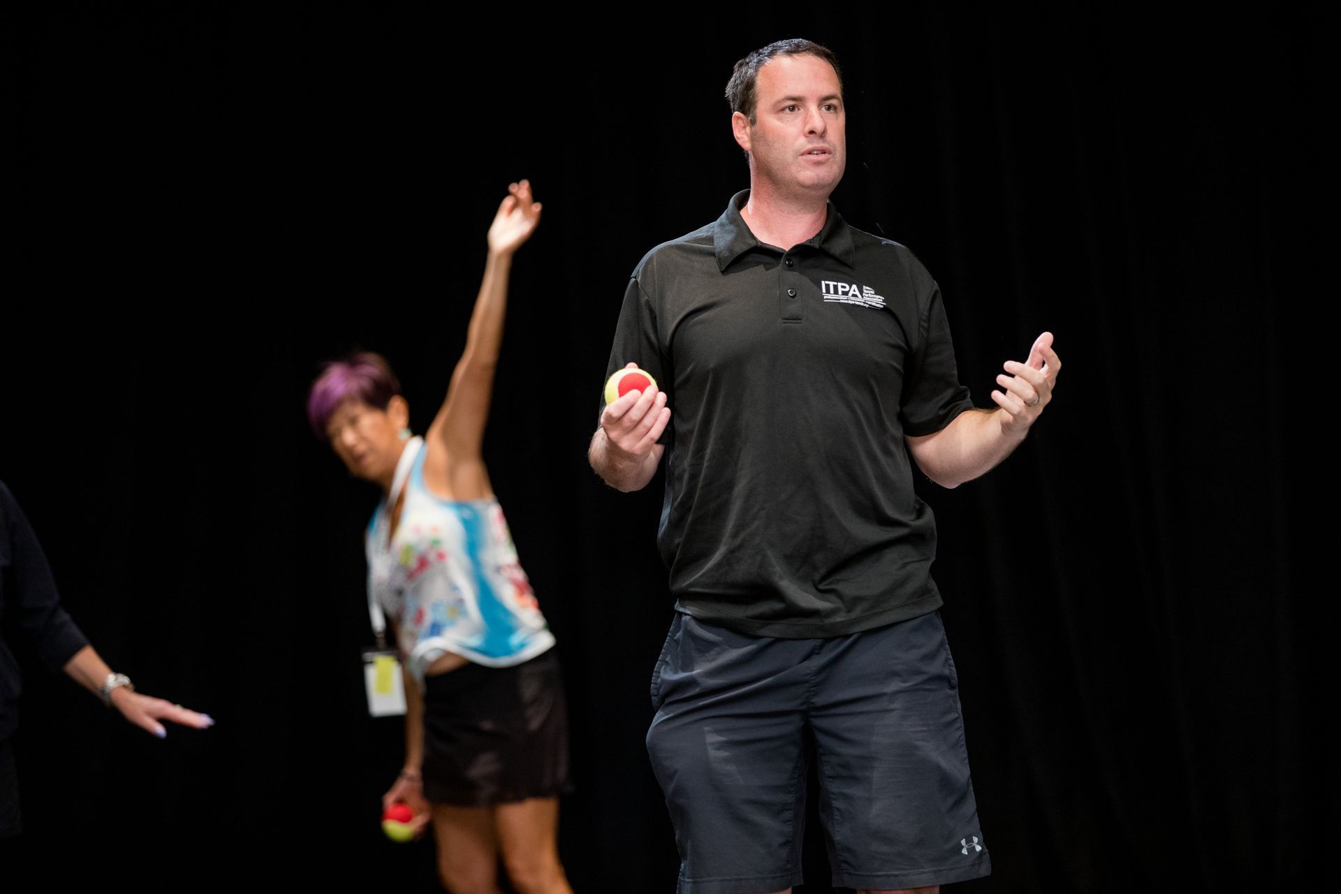Man with red balls explains, woman throws, stage, black background.