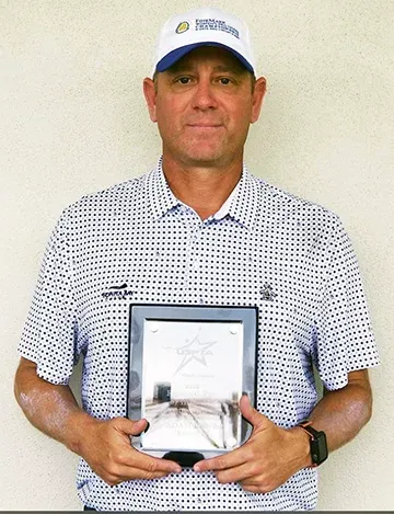 Man in golf attire holds a plaque, smiling, against a light wall. He wears a hat and watch.