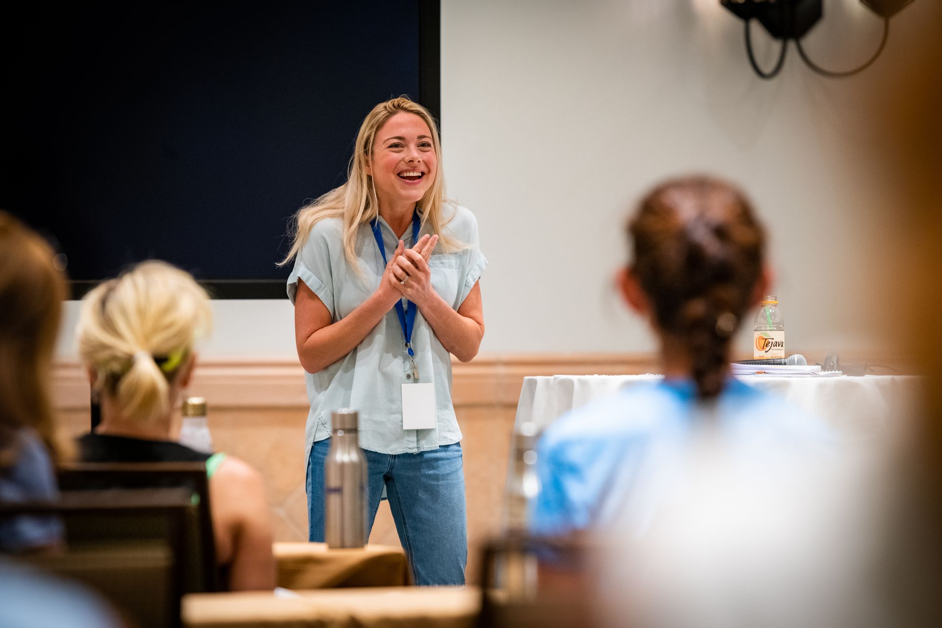 Blonde woman laughing while speaking at a conference. She wears a blue shirt, jeans, and a lanyard. Attendees face her.