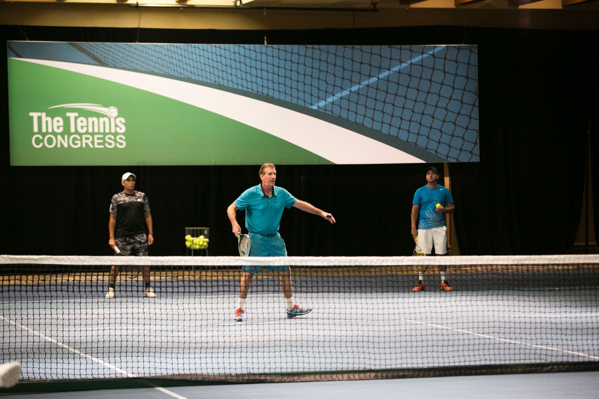 Three people playing tennis indoors. The middle player is about to hit the ball.