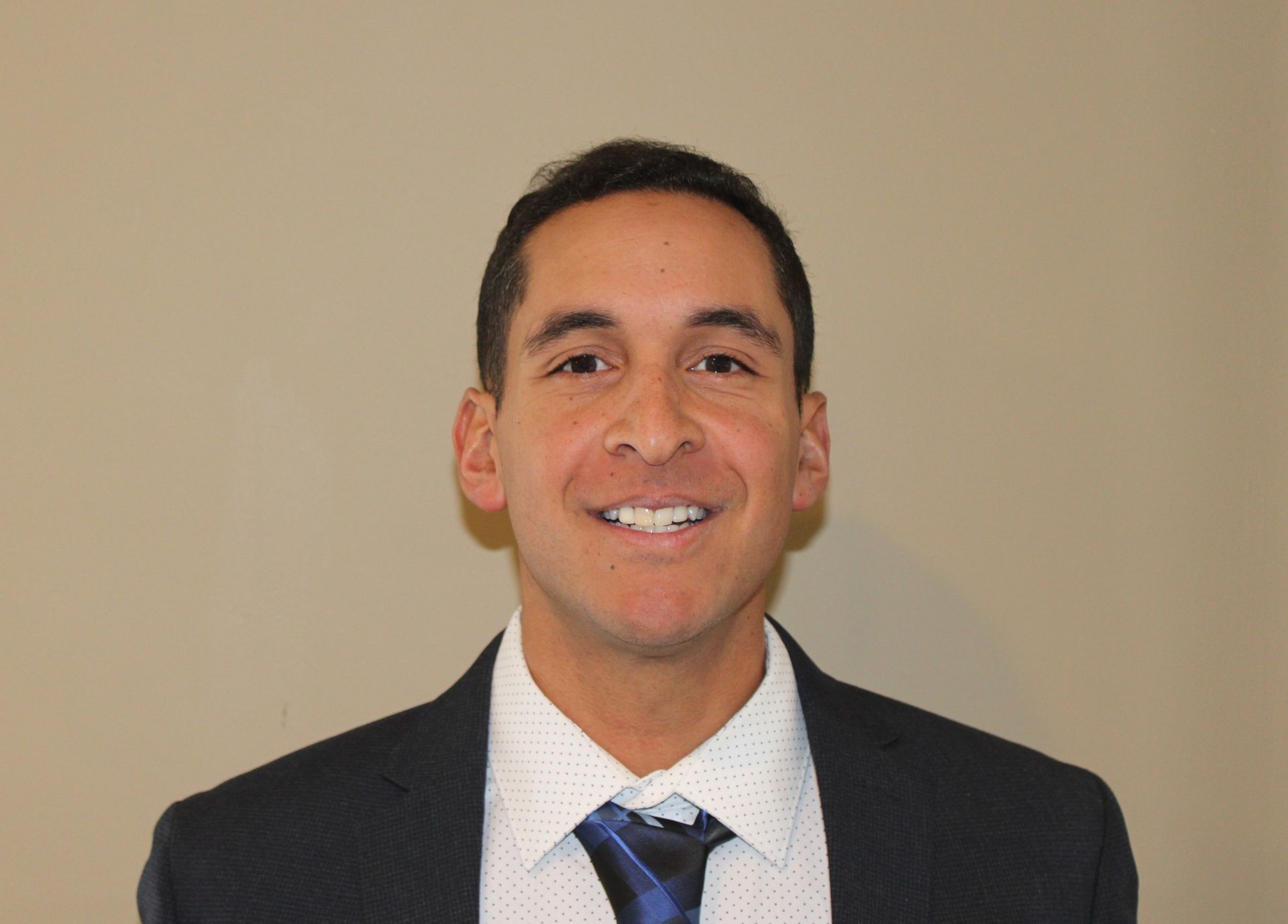 Man in a suit smiles at the camera, against a plain wall.