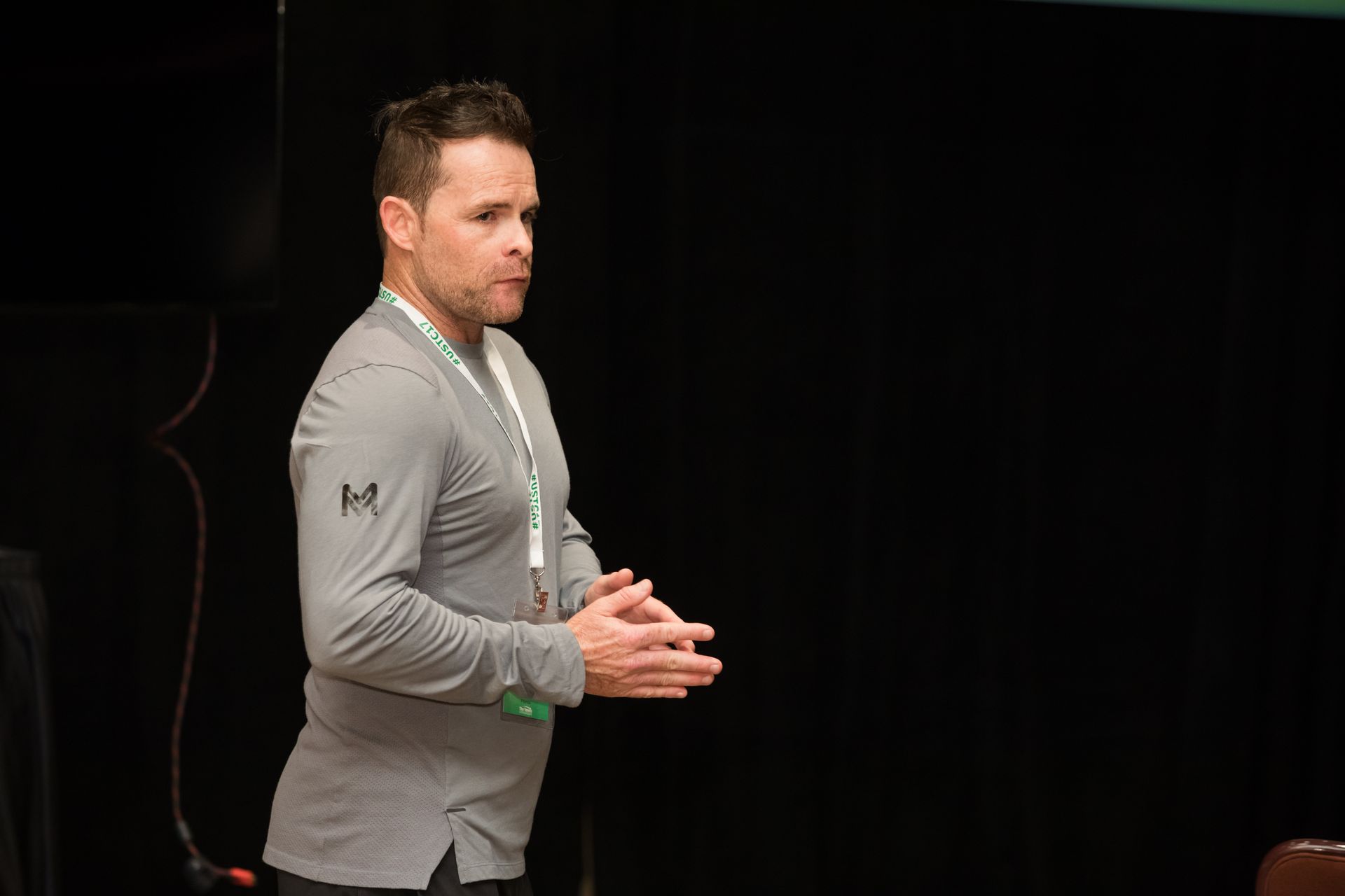 Man in gray shirt speaking, hands clasped, in front of a dark backdrop, wearing a green lanyard.