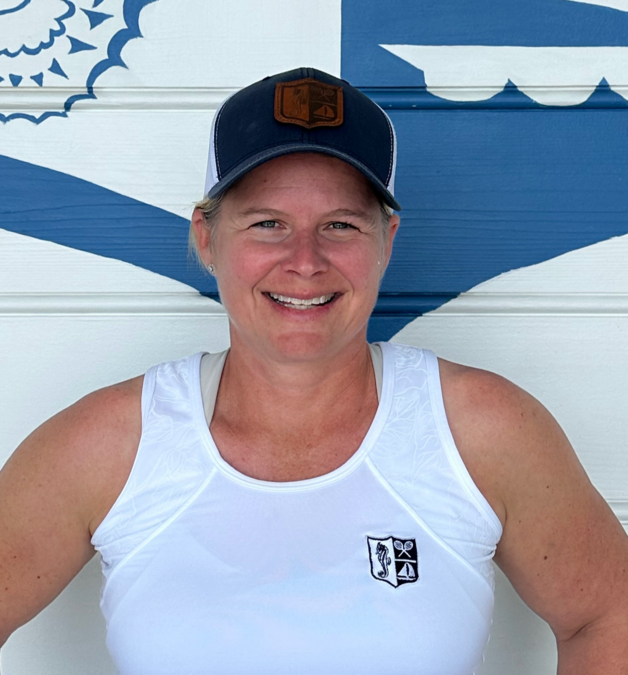Woman wearing a hat and white tank top smiles, standing in front of a blue and white wall.