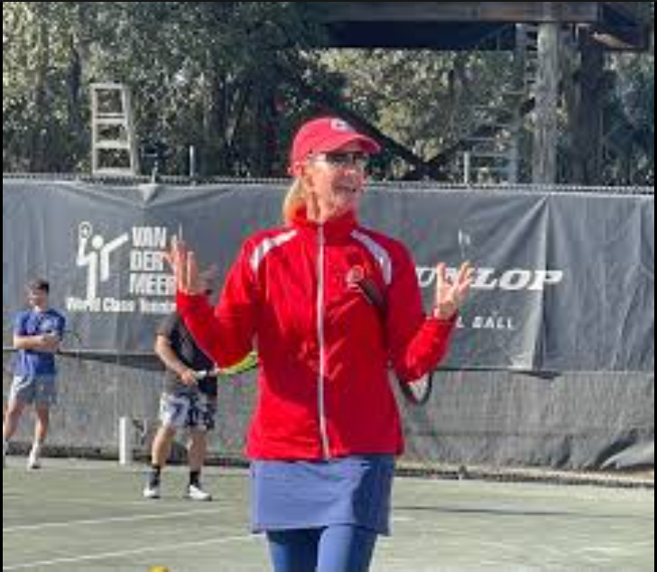 Woman in red jacket and hat on a tennis court, gesturing with hands.
