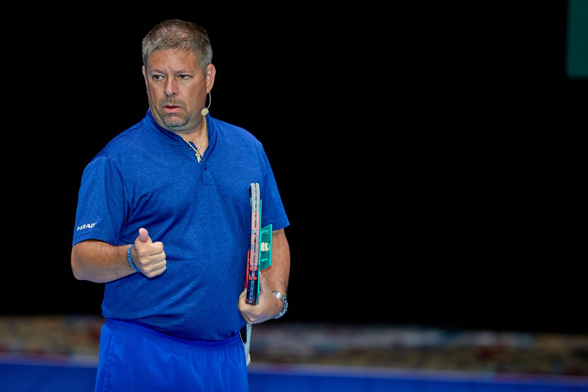 Man in blue polo shirt gives thumbs-up, holding a racket on a court; black background.