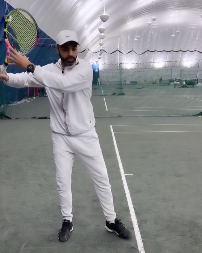 Man in white tennis outfit swings a racquet on indoor court.