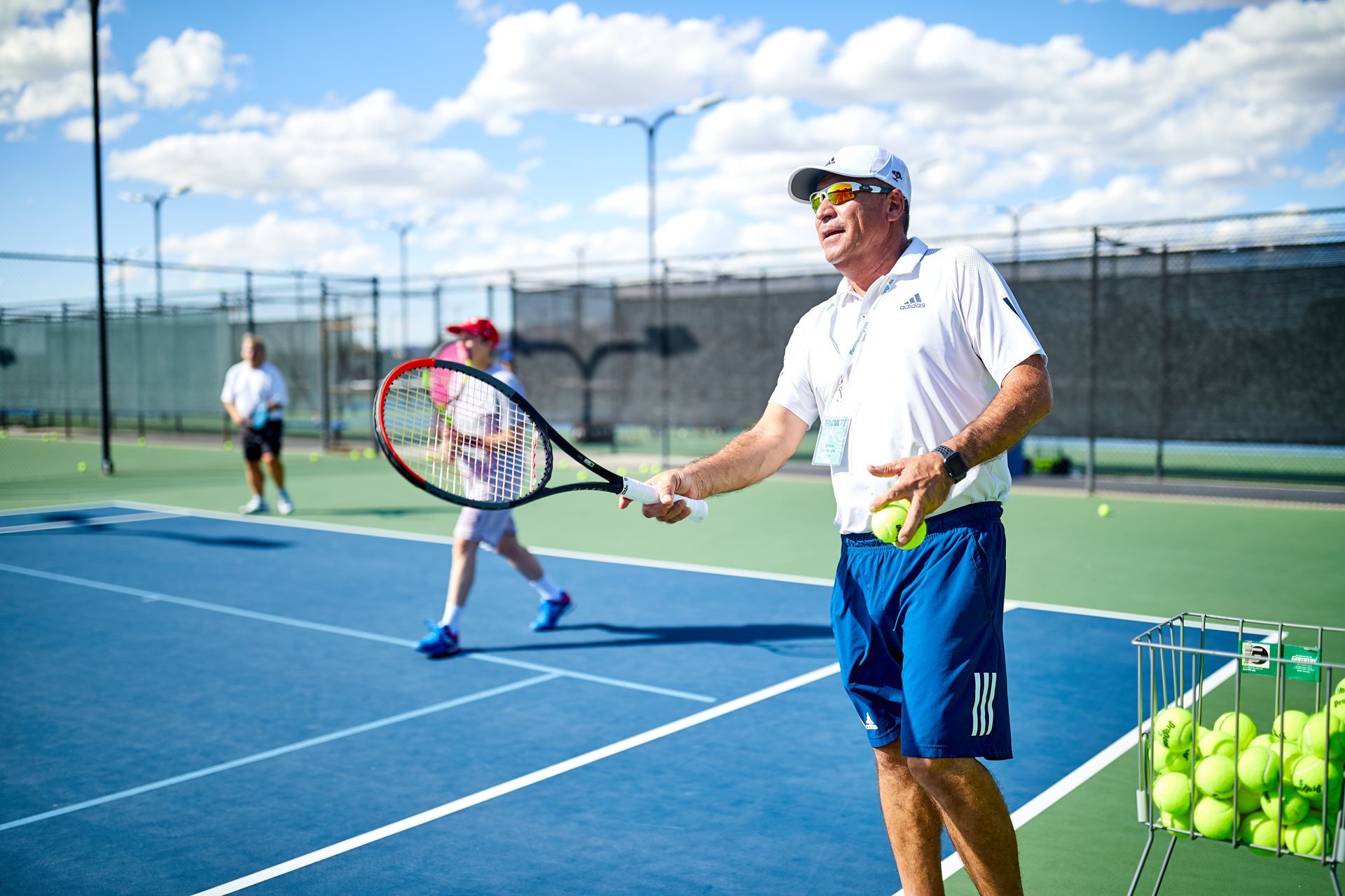 Tennis coach on court, demonstrating a stroke; tennis balls in a basket and players in the background.