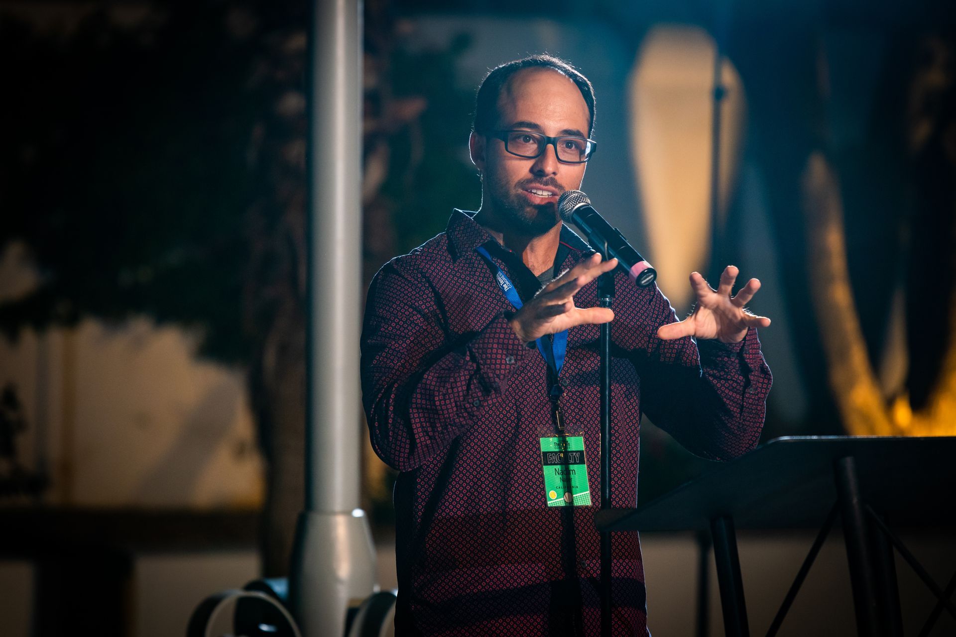 Man with glasses speaks into microphone, gesturing with hands. Outdoor evening setting, wearing patterned shirt.