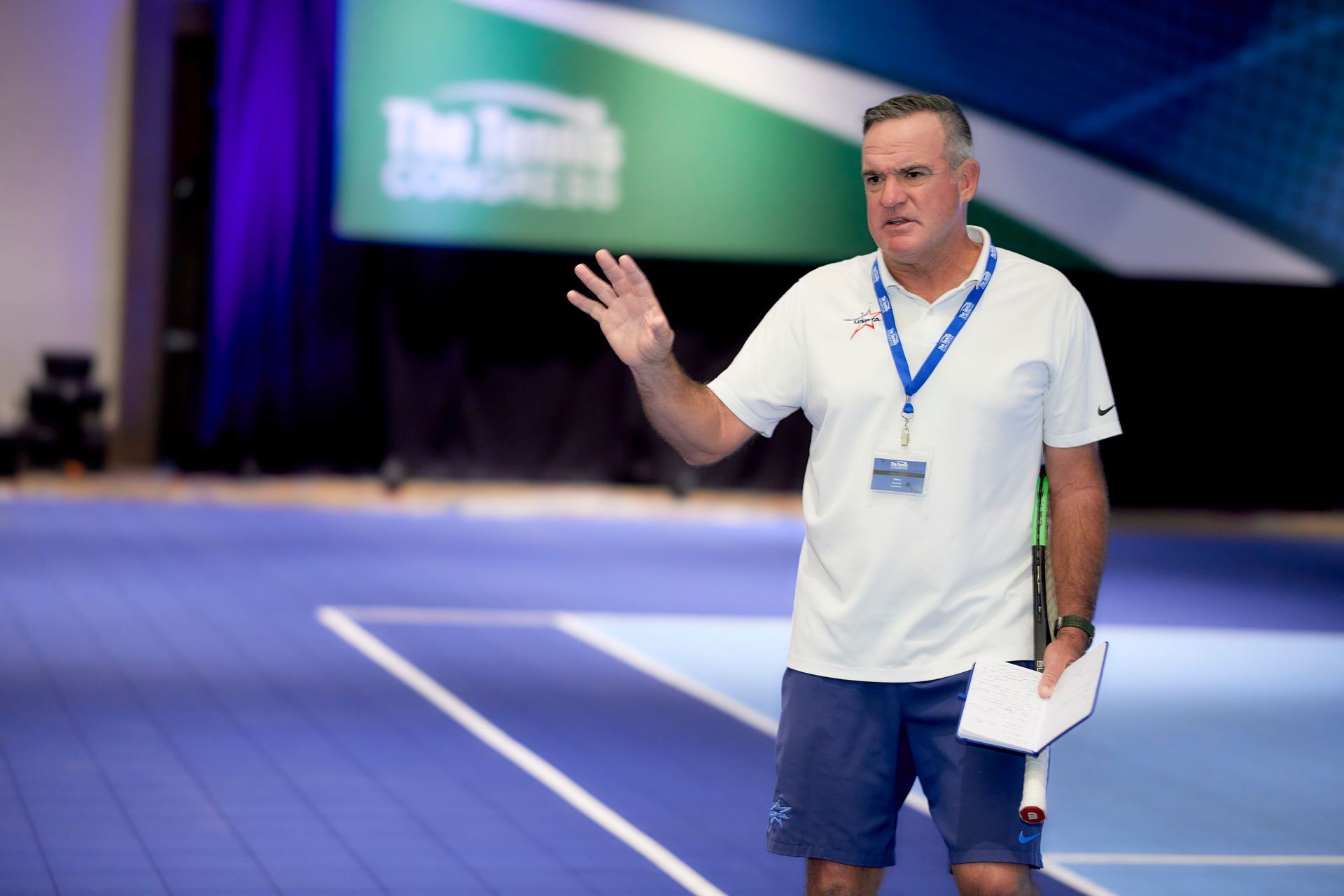 Tennis coach gestures with hand, speaking at a conference. Wearing a white shirt, blue shorts, with blue backdrop.
