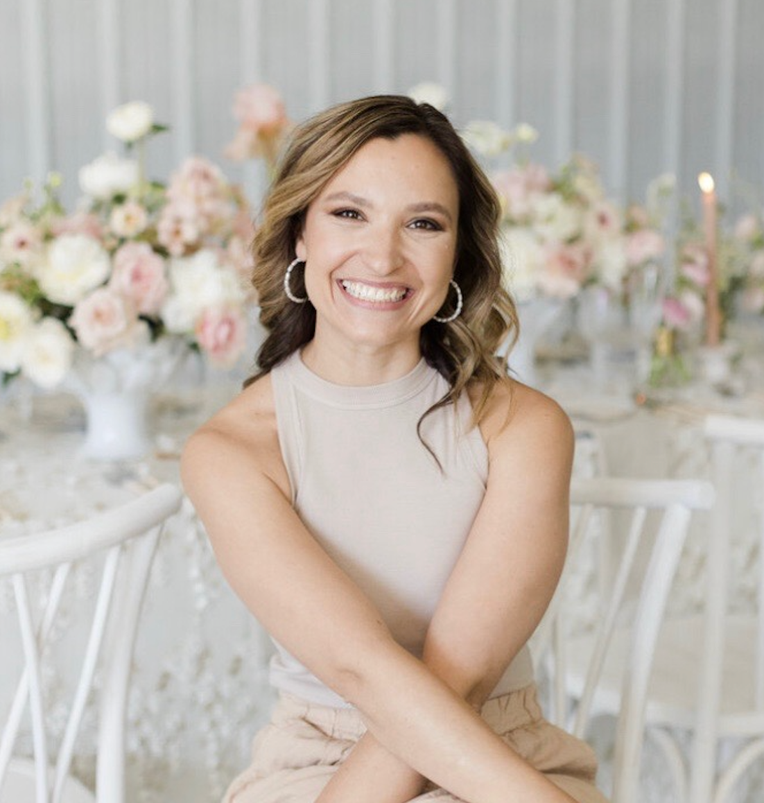 Woman smiling, arms crossed, seated at a table with floral arrangements. Soft colors, indoor setting. Nicole Sirpilla.