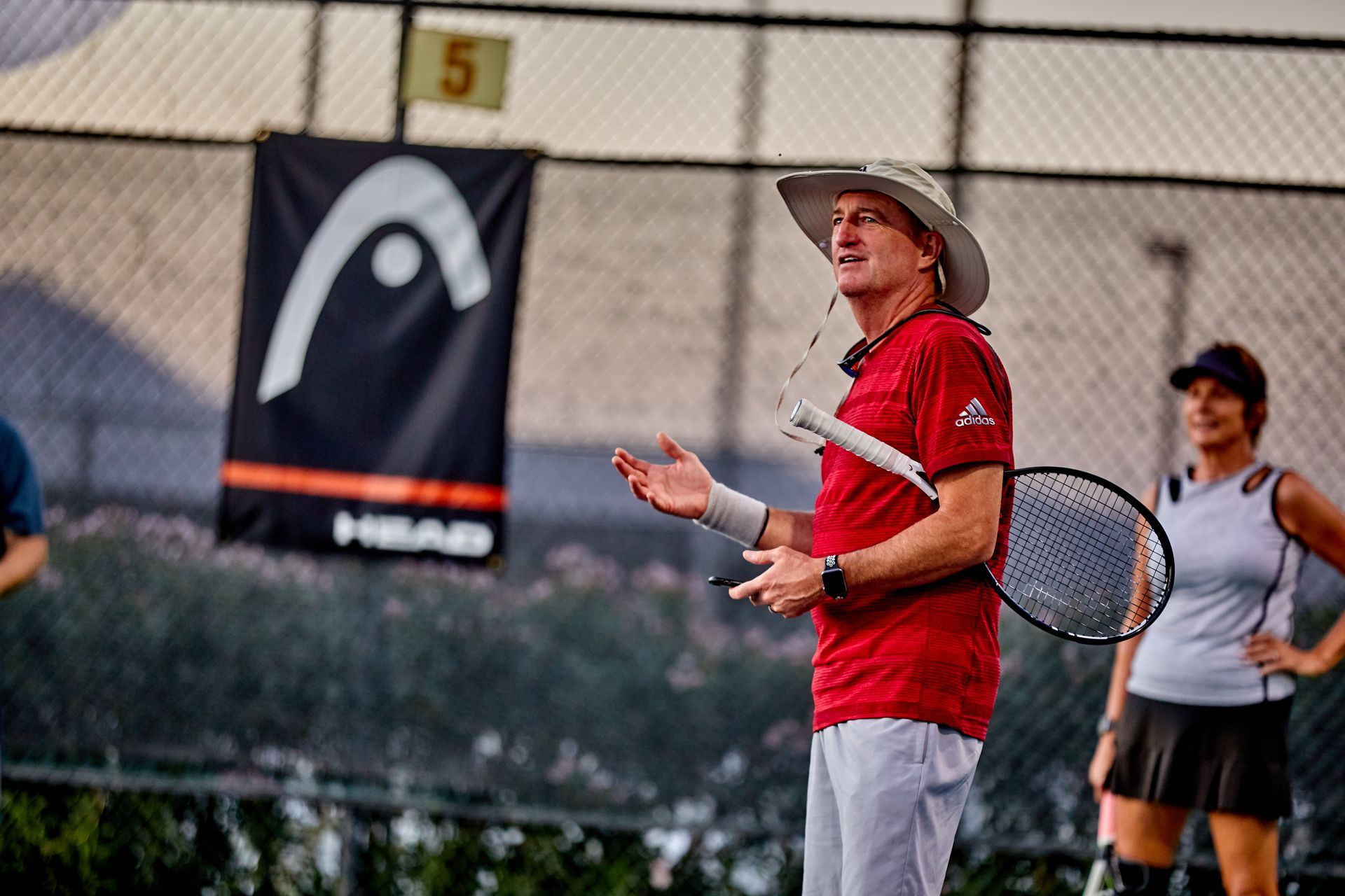 Tennis coach in red shirt, hat, and holding a racquet, talking on a court.