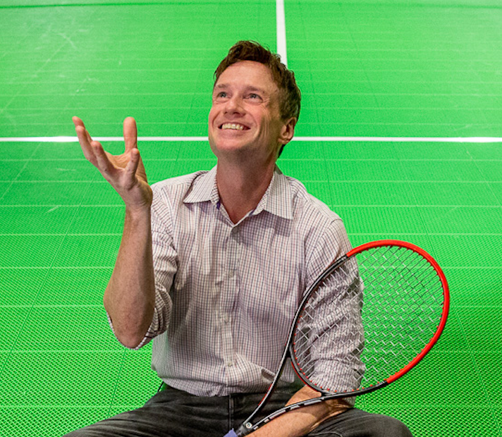 Man smiling, juggling, with tennis racket, on green court.