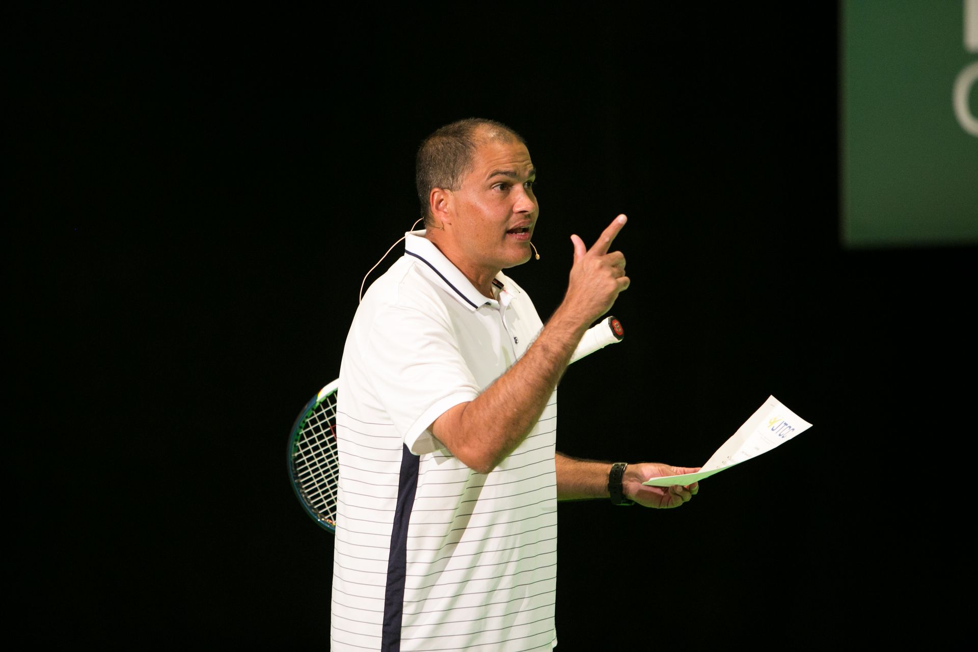 Man with a microphone giving a presentation, holding papers, and pointing. Black background.
