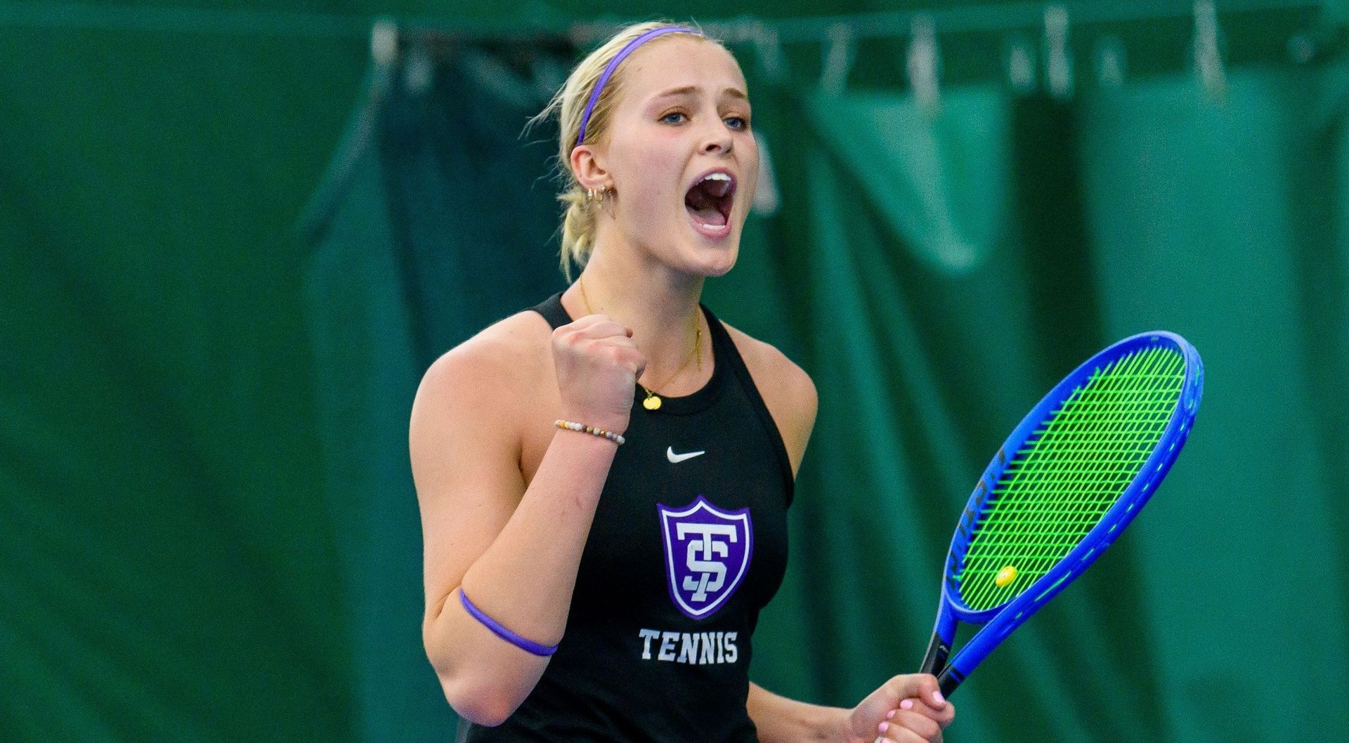 Woman tennis player celebrating a point, wearing a black top with “ST Tennis” on it, inside a green-walled court.