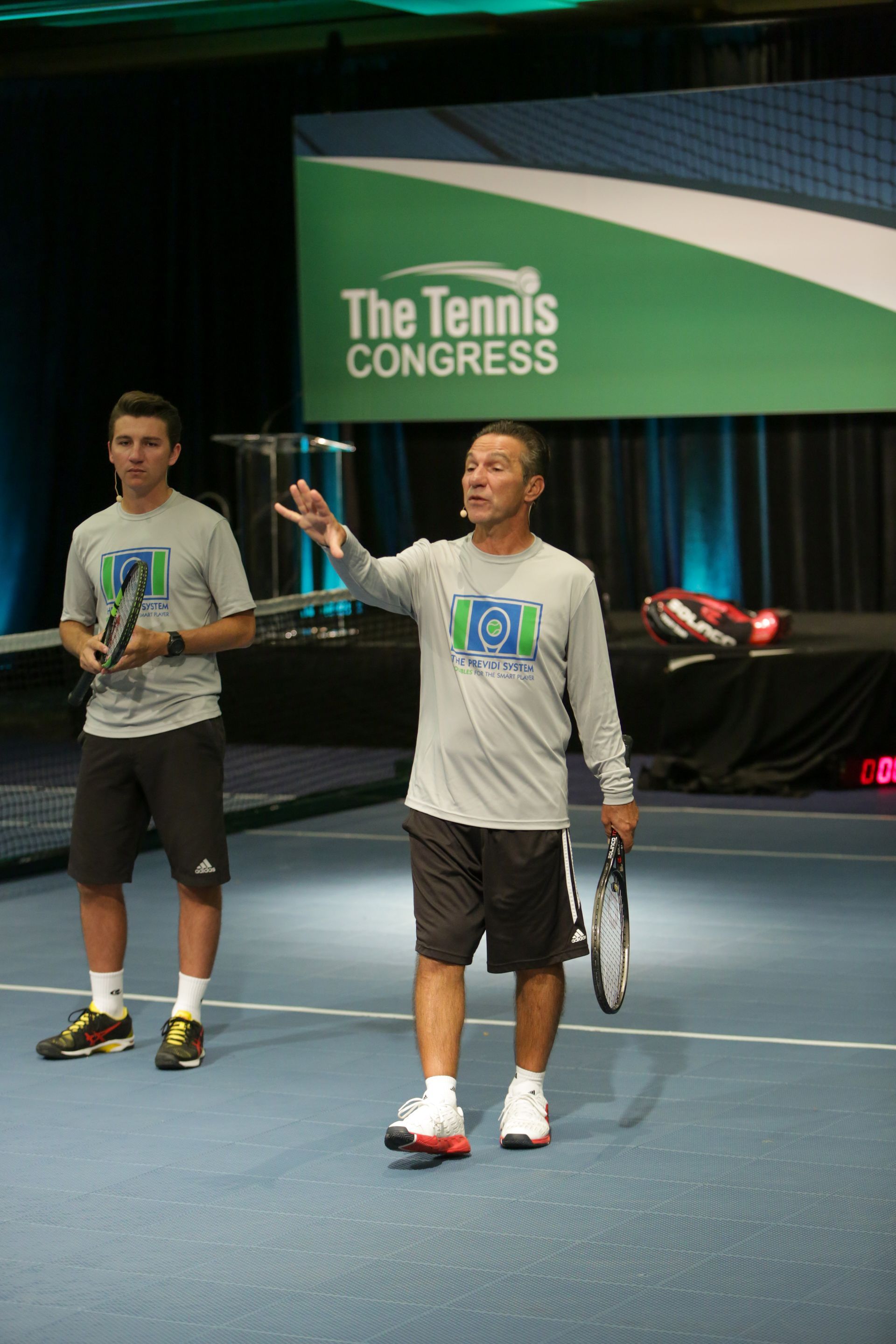 Two men on a tennis court stage; one gestures, the other holds a racket. 