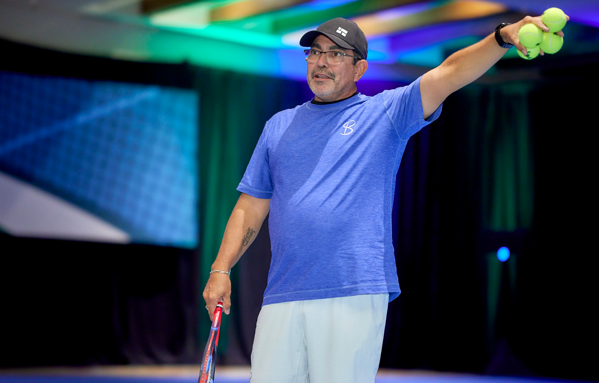Man in blue shirt, holding tennis racket and balls, speaking at a presentation.