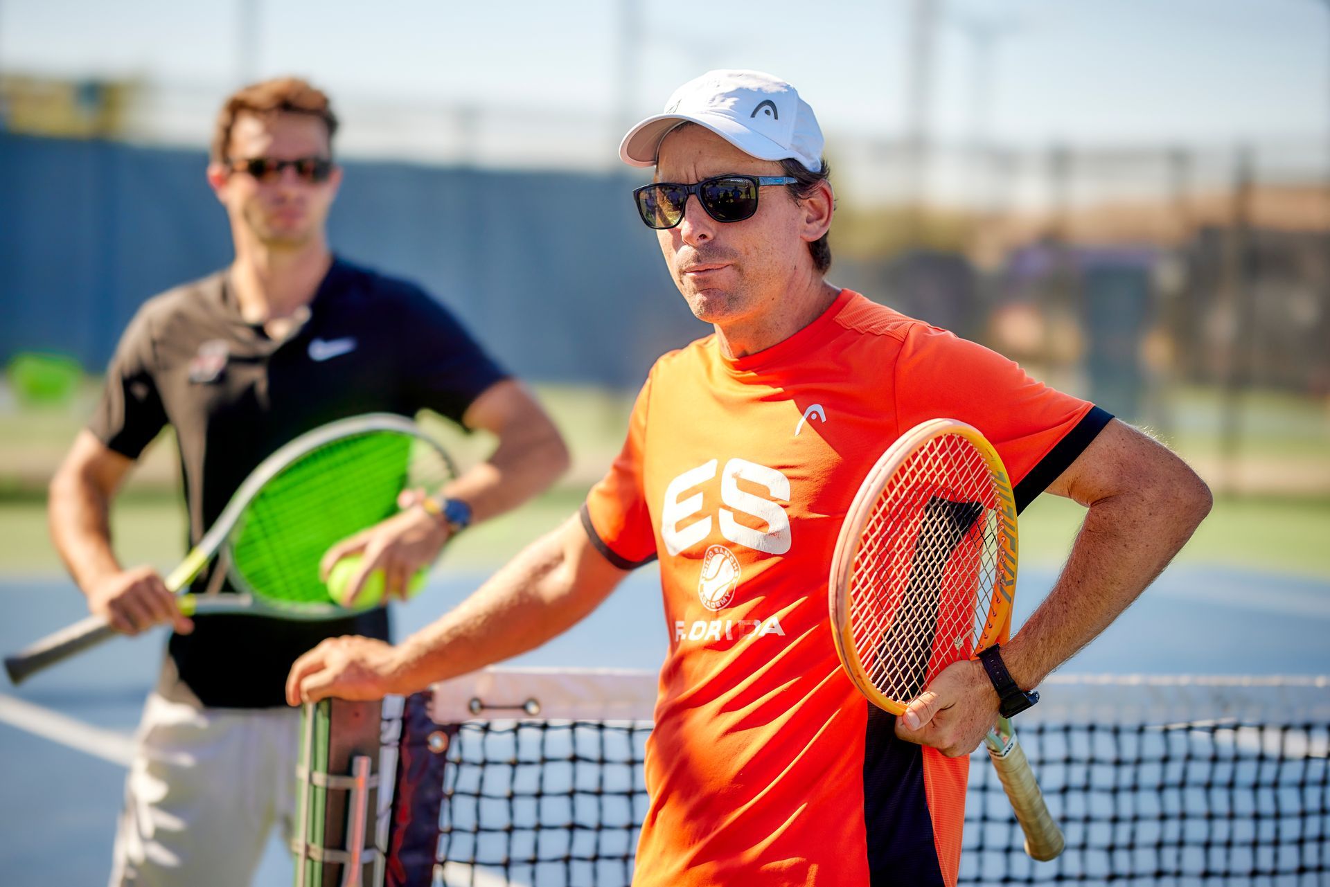 Two men with tennis rackets on a tennis court; one wearing orange, other black, both with sunglasses.
