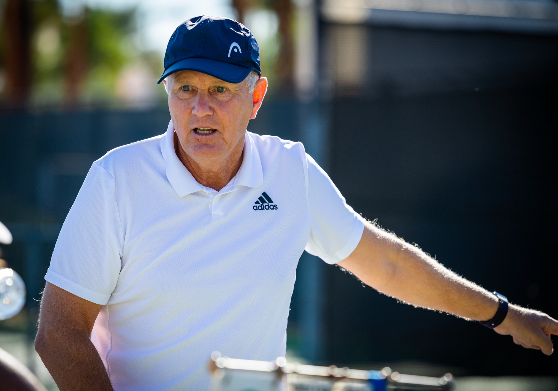 Man in white shirt and blue hat gesturing, outdoors on a tennis court, pointing.