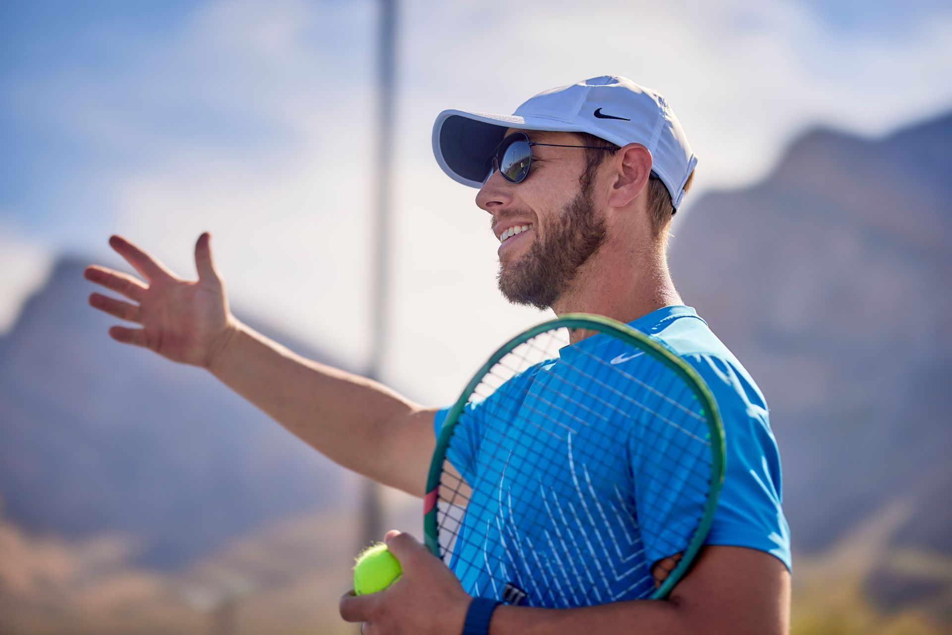 Tennis coach with racket and ball gesturing, smiling outdoors with mountains.