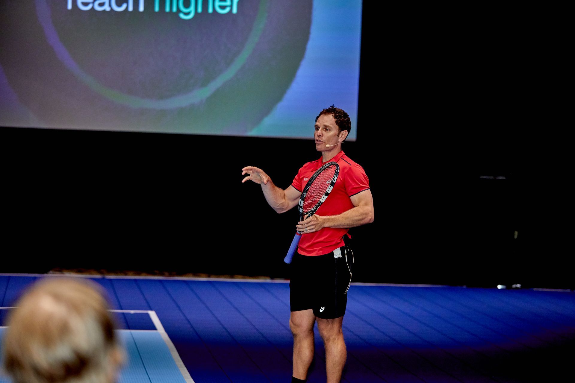 Man with tennis racket gestures while speaking on stage. 
