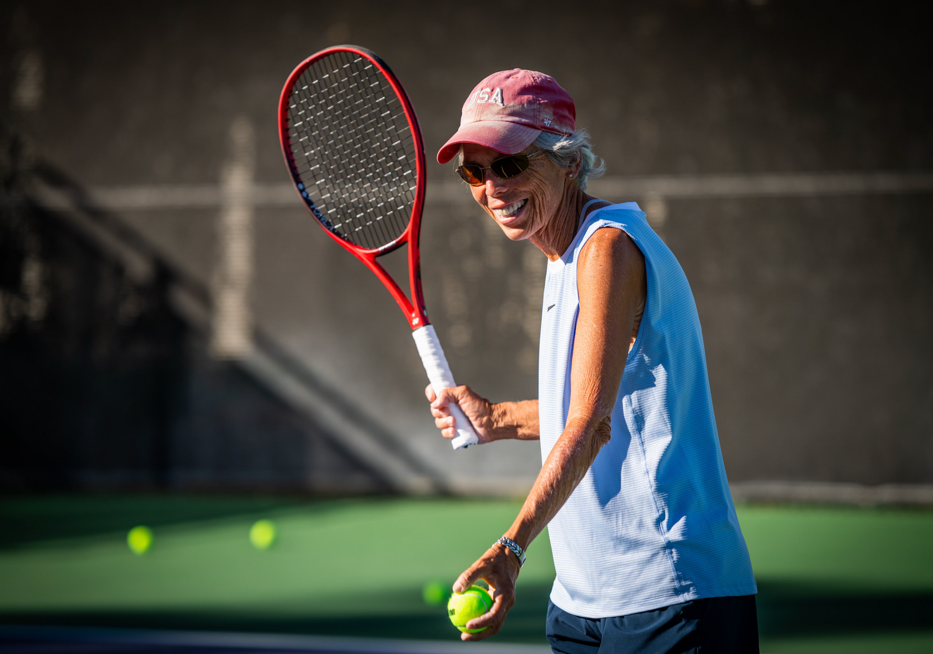 Woman on a tennis court wearing a hat and sunglasses, holding a racket and ball, smiling.