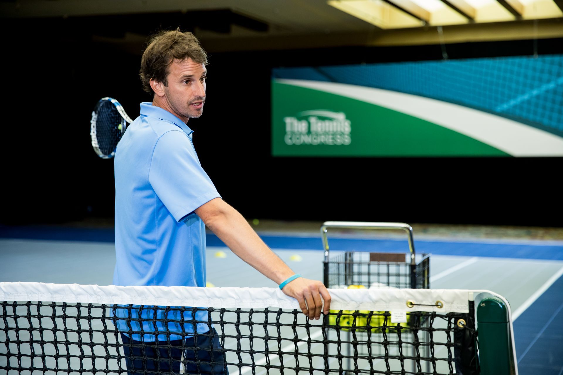 Man in blue shirt leans on tennis net, holding a racket. Tennis court backdrop, 