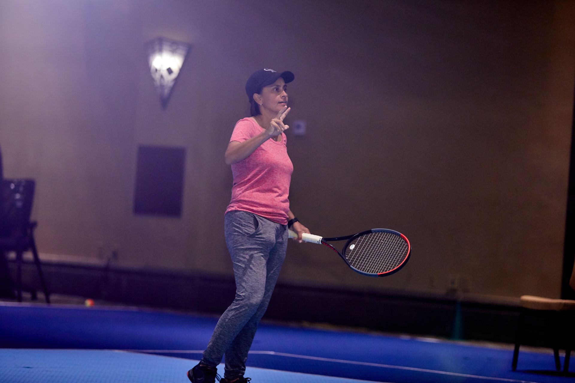 Woman in pink shirt holding tennis racket, gesturing indoors on blue court.