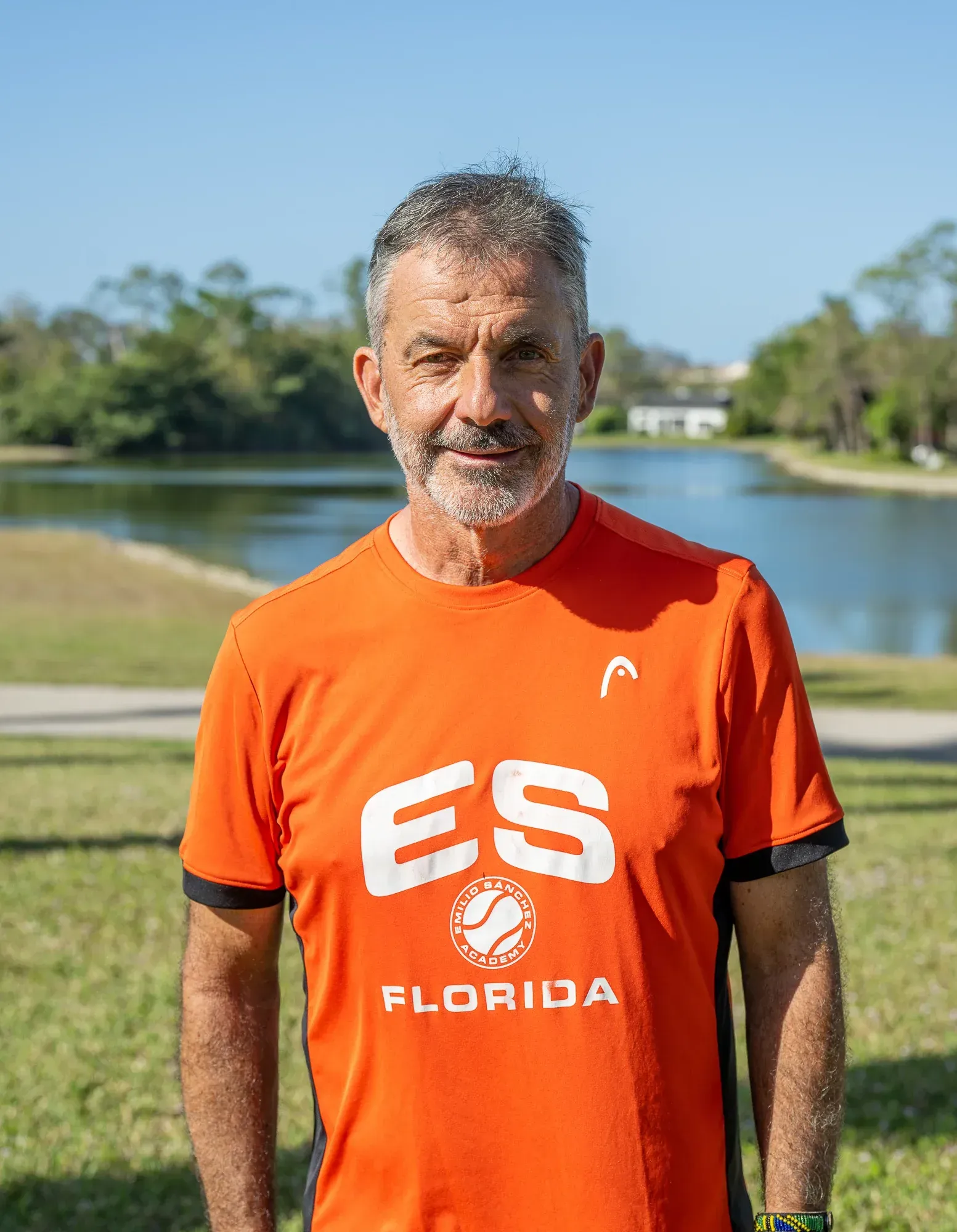 Man in orange tennis shirt stands outside, near a lake.