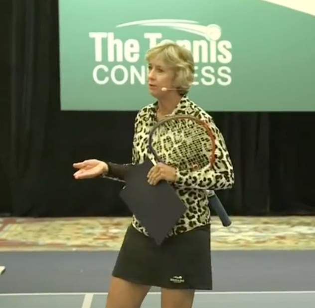 Woman in leopard print shirt holds a tennis racket and clipboard while speaking onstage at a tennis congress.