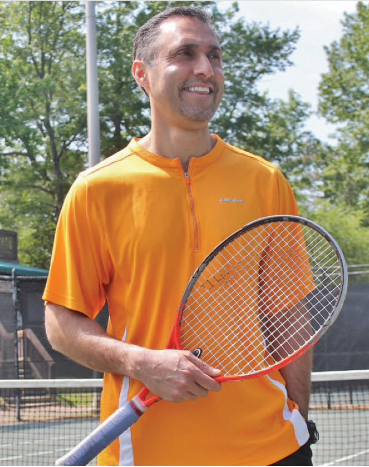 Man in orange shirt holding tennis racket on a tennis court, smiling.