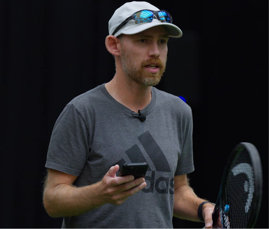 Man in a gray Adidas shirt and cap holding a phone and tennis racket on a dark court