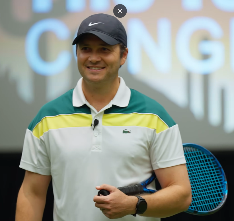 A coach in a white polo shirt stands on a tennis court.