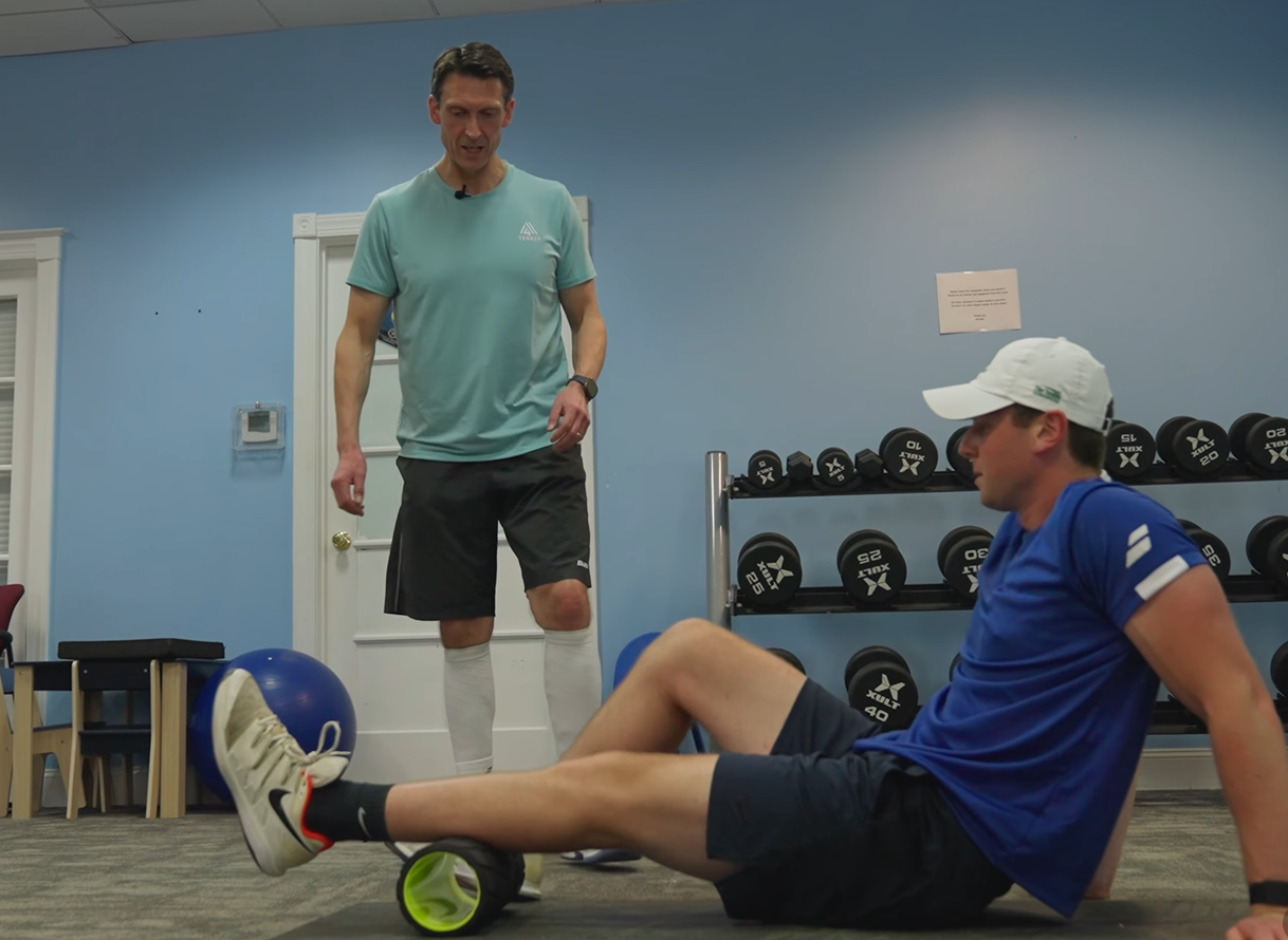 Two men in a gym using foam rollers and a massage ball during a workout recovery session.