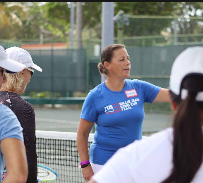 Tennis coach in a blue shirt with her arm extended, talking to a group near a net.